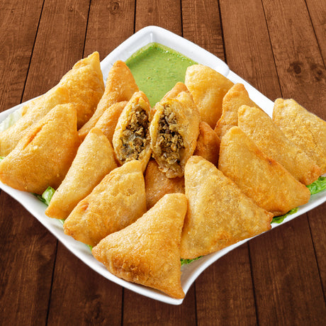 A white square plate filled with golden-brown fried samosas, including two cut open to show the stuffed filling, served with a green chutney on a dark wooden table.