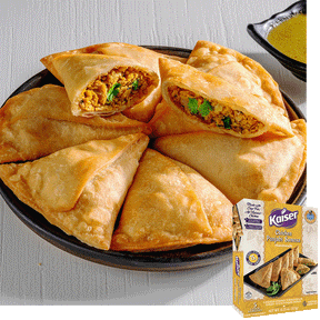 A black plate with 7 chicken punjabi samosas on it arranged into a ring with one samosa on top cut open exposing a a minced chicken stuffing and visible cilantro. A package of Kaiser Chicken Punjabi Samosas is overlaid in the bottom right corner.
