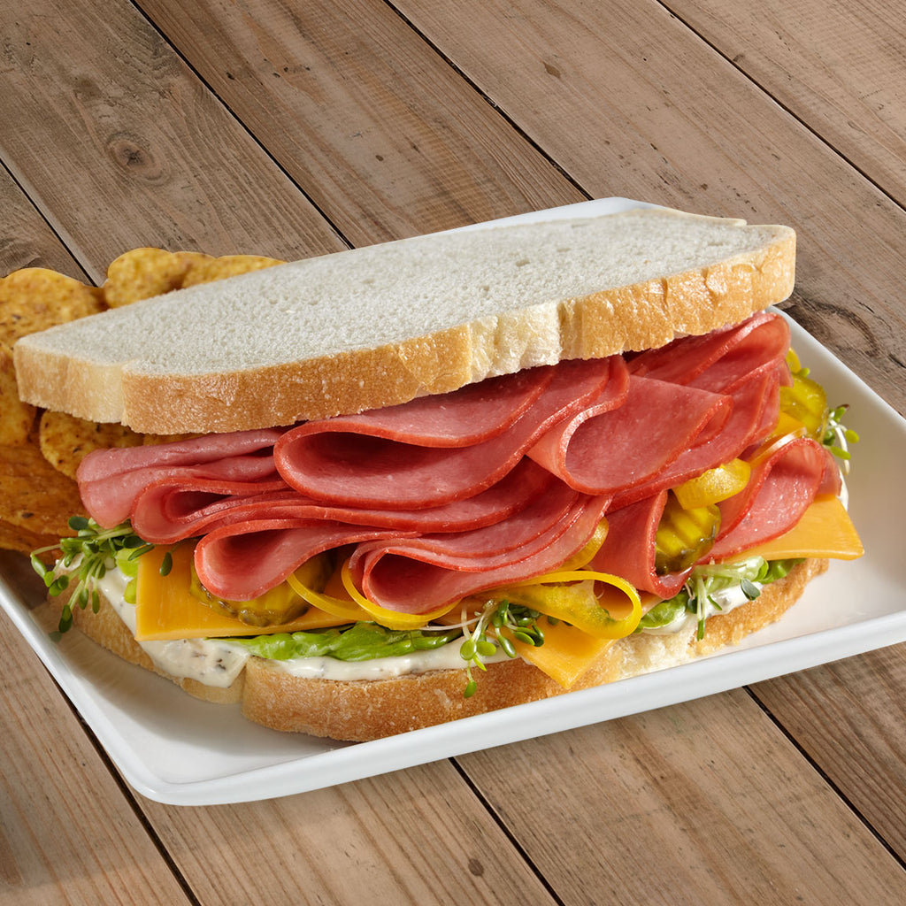 a chicken bologna sandwich on white bread. The chicken bologna is piled high on a bed of American cheese, alfalfa sprouts, lettuce, and a white condiment. At the back of the plate is a small pile of chips. 