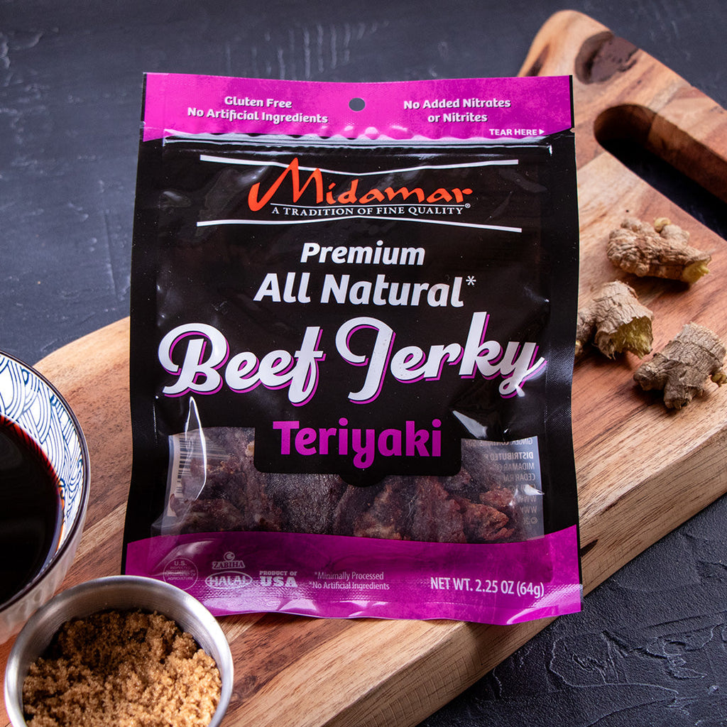 A bag of Midamar Halal Beef jerky Teriyaki sit on a cutting board, a bowls of brown sugar and soy sauce are in the bottom left and piece of ginger are behind the bag. 