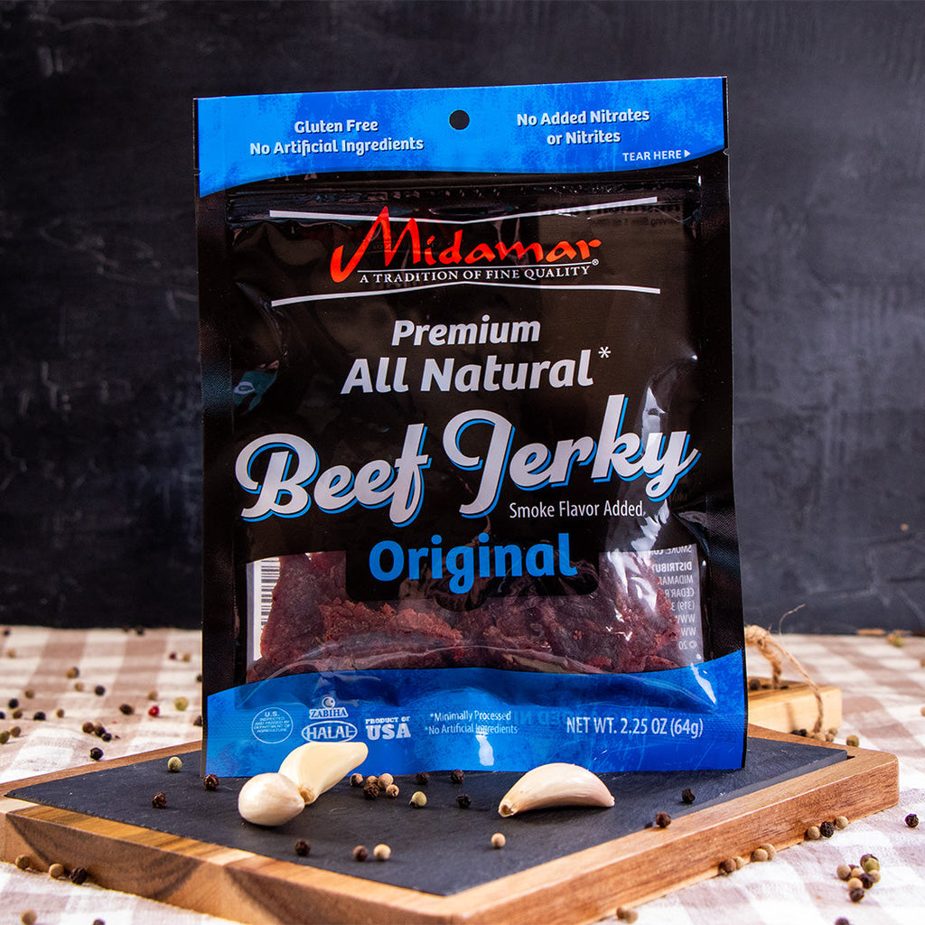 A bag of Midamar Halal Beef Jerky Original is standing on a cutting board and in front of it is whole peppercorns and three cloves of garlic.